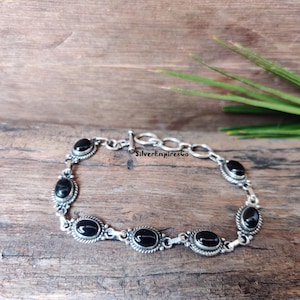 Könnte beinhalten: Ein silbernes Armband mit schwarzen ovalen Edelsteinen. Das Armband hat einen Toggle-Verschluss und ist aus einem Kettenglied-Design gefertigt. Das Armband liegt auf einer Holzoberfläche.