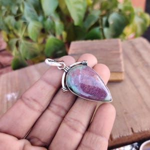 Peut inclure: Un pendentif en argent avec une pierre précieuse verte et rose en forme de larme et une bélière en argent. Le pendentif est tenu dans une main.