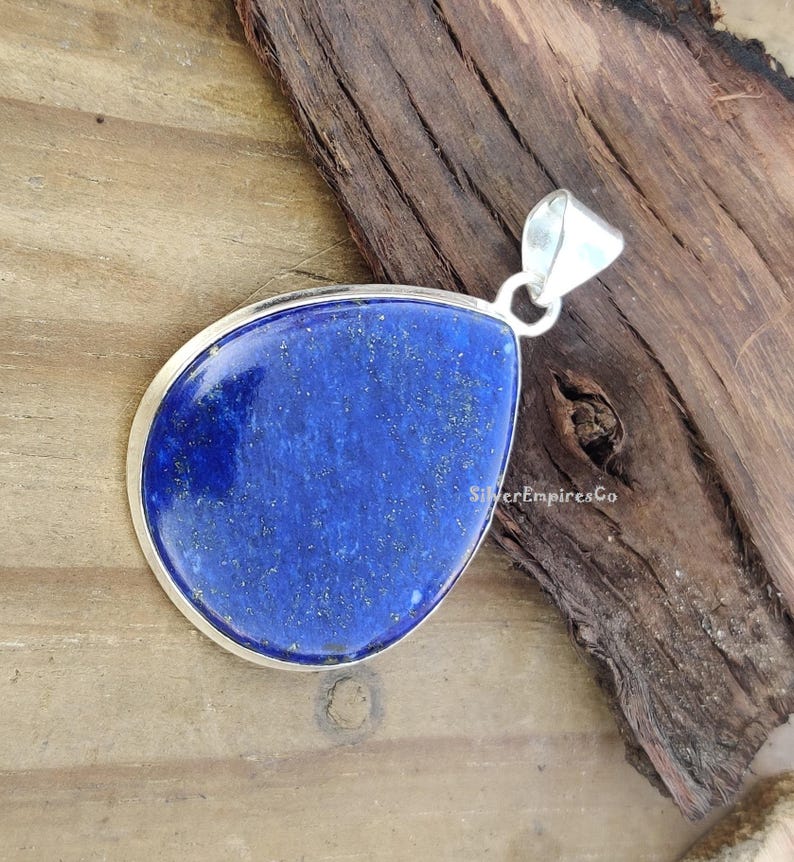 May include: A silver pendant featuring a large, teardrop-shaped lapis lazuli stone. The stone is a deep blue colour with gold flecks throughout. The pendant is displayed on a wooden surface.