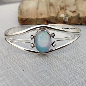 Könnte beinhalten: Ein silberfarbenes Armreif-Armband mit einem ovalen, irisierenden Edelstein in der Mitte. Das Armband hat dekorative Wirbel und den Text "SilverEmpiresCo". Der Edelstein zeigt eine Reihe von Farben, darunter Blau und Grün.