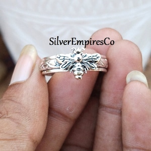 May include: A silver ring featuring a detailed bee design. The ring has a textured band with floral accents and the bee is the focal point. The text "SilverEmpiresCo" is visible in the image.