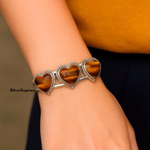 May include: A silver bracelet featuring three heart-shaped tiger's eye stones. The bracelet has a simple, elegant design with a beaded border around each heart. The warm brown and gold tones of the stones contrast with the silver setting.