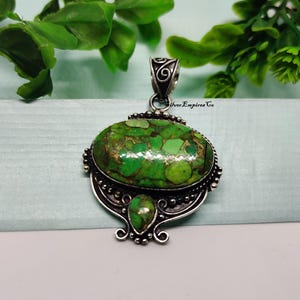 May include: Silver pendant with an oval, green stone with brown and gold accents. The pendant has a decorative silver bail and a smaller teardrop-shaped stone below the main stone. The pendant is set against a light blue background with green foliage.