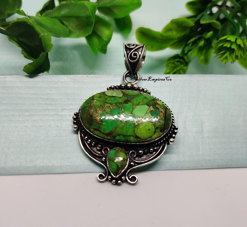 May include: Silver pendant with an oval, green stone with brown and gold accents. The pendant has a decorative silver bail and a smaller teardrop-shaped stone below the main stone. The pendant is set against a light blue background with green foliage.