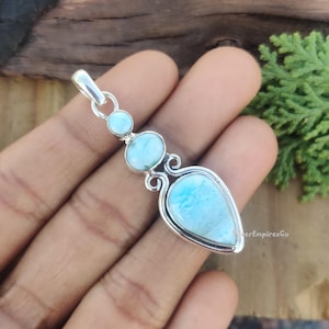 Könnte beinhalten: Ein silberner Anhänger mit drei blauen und weißen Edelstein-Akzenten. Der Anhänger ist tropfenförmig und hat ein zartes, verziertes Design.