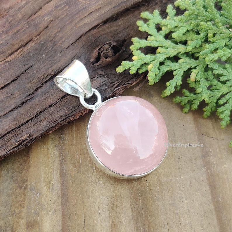 May include: A silver pendant with a round, light pink rose quartz gemstone. The pendant is on a wooden surface.