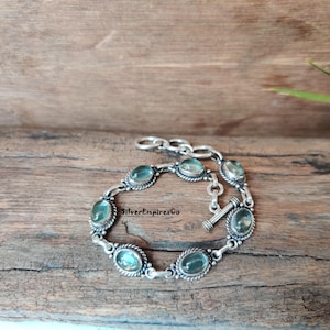 Peut inclure: Un bracelet en argent orné de pierres précieuses ovales bleu clair. Le bracelet est doté d'un fermoir à bascule et est présenté sur une surface en bois. Le texte "SilverEmpiresCo" est visible.