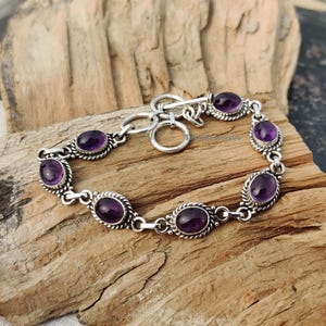 May include: A silver bracelet featuring oval-shaped, deep purple gemstones. The gemstones are set in a decorative silver frame and linked together with a chain. The bracelet is displayed on a wooden surface.