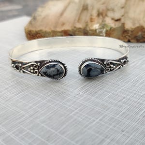 Könnte beinhalten: Ein silbernes Armreif mit zwei tropfenförmigen schwarzen und weißen Steinen. Das Armband hat ein zartes Blumenmuster.