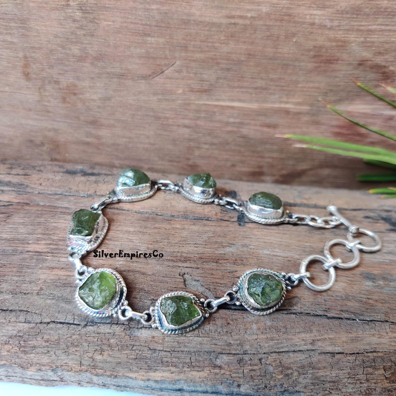 May include: A silver bracelet with a toggle clasp features seven green gemstone beads. The beads are irregularly shaped and have a rough, natural texture. The bracelet is on a wooden surface.