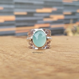 Puede incluir: Un anillo de plata con textura martillada y una piedra preciosa ovalada azul claro. El anillo tiene una banda ancha y un acabado pulido, que muestra el color de la piedra preciosa. El anillo se exhibe sobre una superficie de madera.