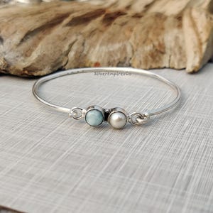 Puede incluir: Un brazalete de plata con una piedra preciosa azul claro y una perla blanca. El brazalete está sobre una superficie gris claro.