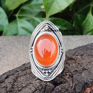 Peut inclure: Une bague en argent avec une pierre de cornaline orange sertie dans une bélière ovale ornée. La bague a une bande texturée et des détails complexes.