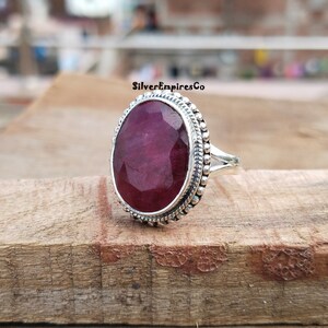Puede incluir: Un anillo de plata con una piedra preciosa ovalada, facetada y de color rojo intenso. La piedra preciosa est&aacute; engastada en un bisel detallado con cuentas. La banda del anillo tiene un dise&ntilde;o dividido. El texto "SilverEmpiresCo" es visible en el fondo.