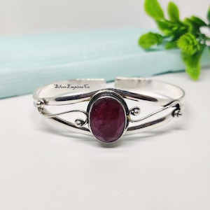 Könnte beinhalten: Ein silbernes Armreif-Armband mit einem ovalen, tiefroten Edelstein in der Mitte. Das Armband hat ein dekoratives, fließendes Design mit kleinen, runden Akzenten. Der Text "Silver Empires Co" ist sichtbar.