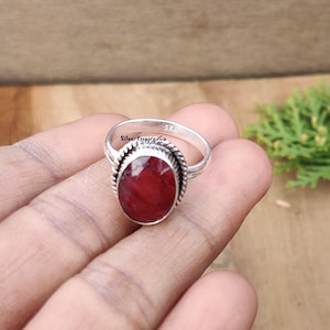 Könnte beinhalten: Ein silberner Ring mit einem roten ovalen Edelstein, der in einer silbernen Fassung sitzt. Der Ring befindet sich auf einer Hand.