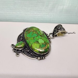 May include: A silver pendant with a large, oval-shaped green stone and a smaller, teardrop-shaped stone. The stones have a mottled appearance with shades of green and brown. The pendant has an ornate, detailed silver setting.