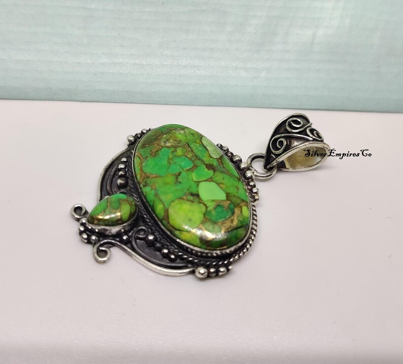May include: A silver pendant with a large, oval-shaped green stone and a smaller, teardrop-shaped stone. The stones have a mottled appearance with shades of green and brown. The pendant has an ornate, detailed silver setting.