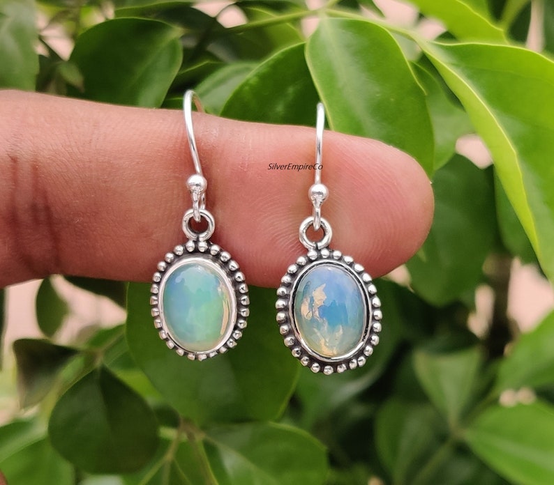 May include: Silver drop earrings featuring oval opal stones. The earrings have a beaded border and a simple hook fastening. The opals display a milky, iridescent quality, with touches of blue and green. The earrings are set against a blurred green backdrop.