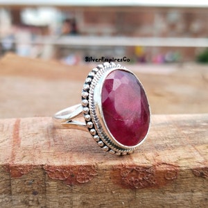 Puede incluir: Un anillo de plata con una gran gema de rub&iacute; facetada de forma ovalada. El rub&iacute; es de color rojo intenso y est&aacute; rodeado por un borde plateado con cuentas. El anillo est&aacute; sobre una superficie de madera, con el texto "SilverEmpiresCo".