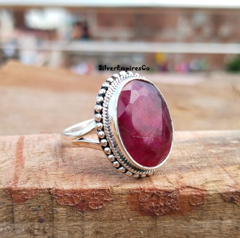 Puede incluir: Un anillo de plata con una gran gema de rub&iacute; facetada de forma ovalada. El rub&iacute; es de color rojo intenso y est&aacute; rodeado por un borde plateado con cuentas. El anillo est&aacute; sobre una superficie de madera, con el texto "SilverEmpiresCo".