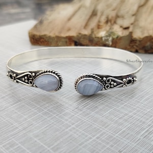 Könnte beinhalten: Ein silbernes Armreif-Armband mit zwei blauen Achat-Steinen, die vorne eingesetzt sind. Das Armband hat ein zartes Blumenmuster.