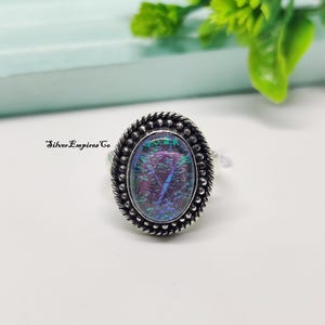 Könnte beinhalten: Ovaler Silberring mit einem schimmernden, irisierenden Stein in Lila, Blau und Grün. Der Ring hat einen strukturierten, perlenbesetzten Rand und den Text "Silver Empires Co".
