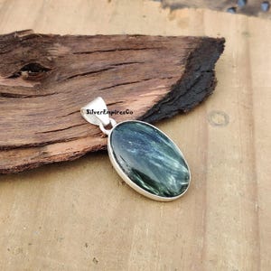 Op de afbeelding: Een zilveren hanger met een ovale, groen-witte edelsteen. De hanger ligt op een houten oppervlak. De tekst "SilverEmpiresCo" is zichtbaar op de hanger.