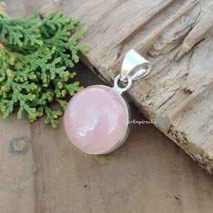 May include: A round, pale pink rose quartz pendant with a silver bail. The pendant is on a wooden surface.