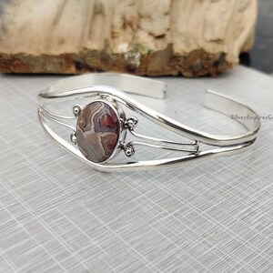 May include: A silver cuff bracelet featuring an oval stone with a pattern in brown, grey, and red. The bracelet has a decorative design with small silver accents around the stone. The open design allows for easy wear.