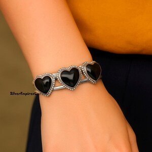 May include: Silver cuff bracelet featuring three black heart-shaped stones. Each heart is outlined with small, silver-colored beads. The bracelet is worn on a wrist against a yellow and navy blue background.