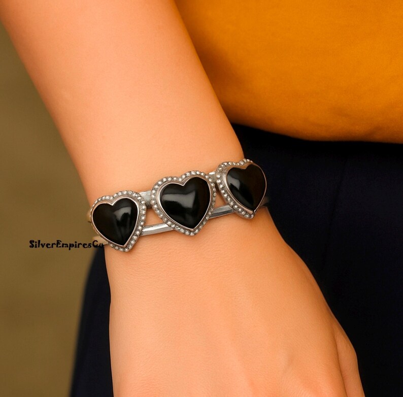 May include: Silver cuff bracelet featuring three black heart-shaped stones. Each heart is outlined with small, silver-colored beads. The bracelet is worn on a wrist against a yellow and navy blue background.