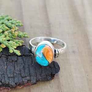 Puede incluir: Anillo de plata con una piedra ovalada turquesa y naranja. El anillo tiene una banda de plata pulida y un engaste decorativo alrededor de la piedra. Los colores de la piedra contrastan con la plata.