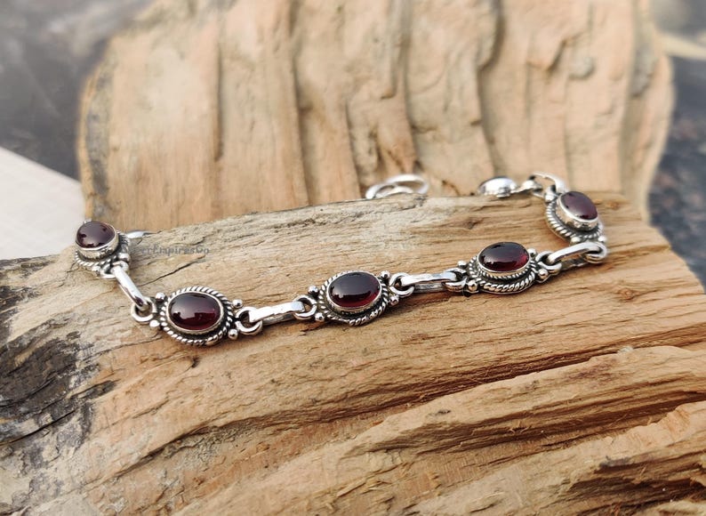 May include: A silver bracelet featuring five oval-shaped red gemstones. The bracelet has a delicate chain design.