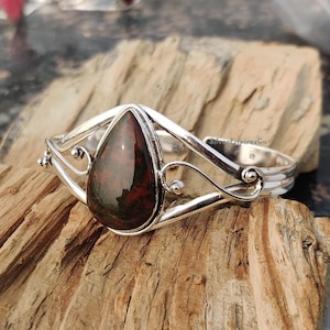 Könnte beinhalten: Ein silbernes Armband mit einem tropfenförmigen grünen und roten Edelstein. Das Armband hat ein zartes, verziertes Design.