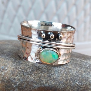 May include: A silver-colored spinner ring with a hammered texture. The ring features an oval-shaped opal stone and three small decorative studs. The text "SilverEmpiresCo" is visible on the ring.