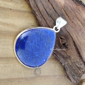 May include: A silver pendant featuring a large, teardrop-shaped lapis lazuli stone. The stone is a deep blue colour with gold flecks throughout. The pendant is displayed on a wooden surface.