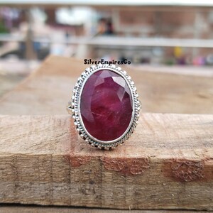 Puede incluir: Anillo de plata con una gran gema de rub&iacute; de corte ovalado. El rub&iacute; es de un color rojo intenso y rico. El anillo tiene un borde decorativo con cuentas. El anillo est&aacute; sobre una superficie de madera.
