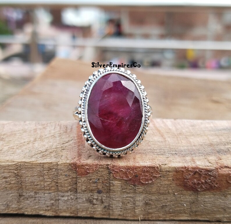Puede incluir: Anillo de plata con una gran gema de rub&iacute; de corte ovalado. El rub&iacute; es de un color rojo intenso y rico. El anillo tiene un borde decorativo con cuentas. El anillo est&aacute; sobre una superficie de madera.