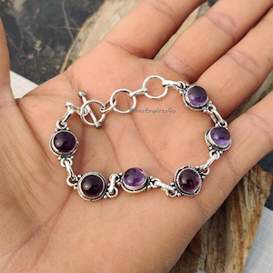 Könnte beinhalten: Ein silbernes Armband mit einem Schiebeverschluss und sechs runden Amethyst-Edelsteinen. Das Armband befindet sich am Handgelenk einer Person.