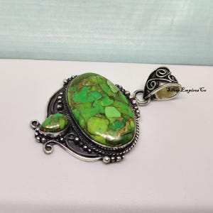 May include: A silver pendant with a large, oval-shaped green stone and a smaller, teardrop-shaped stone. The stones have a mottled appearance with shades of green and brown. The pendant has an ornate, detailed silver setting.