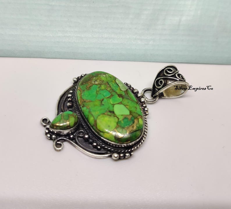 May include: A silver pendant with a large, oval-shaped green stone and a smaller, teardrop-shaped stone. The stones have a mottled appearance with shades of green and brown. The pendant has an ornate, detailed silver setting.
