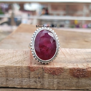 Könnte beinhalten: Silberner Ring mit einem großen, oval geschliffenen Rubin. Der Rubin hat eine satte, tiefrote Farbe. Der Ring hat einen dekorativen, perlenbesetzten Rand. Der Ring liegt auf einer Holzoberfläche.