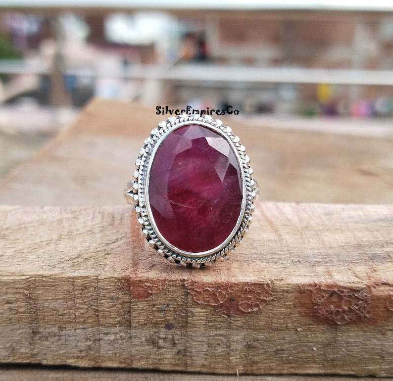 Puede incluir: Anillo de plata con una gran gema de rub&iacute; de corte ovalado. El rub&iacute; es de un color rojo intenso y rico. El anillo tiene un borde decorativo con cuentas. El anillo est&aacute; sobre una superficie de madera.