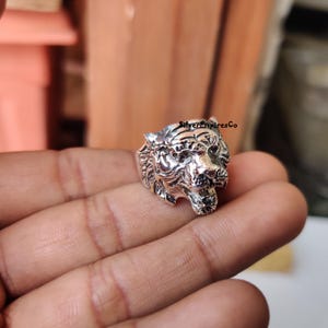 Könnte beinhalten: Ein silberner Ring mit einem detaillierten Tigerkopf-Design. Der Ring wird an einem Finger getragen und der Tiger hat den Mund offen.