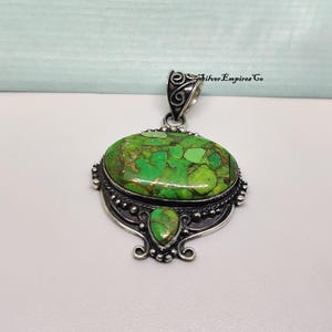 May include: A silver pendant with a large, oval-shaped green stone with brown veining. The pendant has a smaller, teardrop-shaped stone below the oval. The metalwork is ornate, with decorative details and the text "Silver Empires Co."