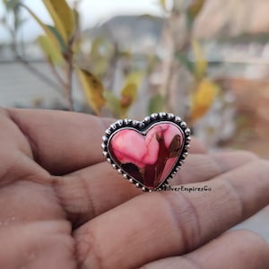 Könnte beinhalten: Ein herzförmiger Ring mit einem rosa und kastanienbraunen Stein, der in einem silbernen Band gefasst ist. Der Rand des Rings ist mit kleinen, runden Silberperlen verziert. Dies ist ein Schmuckstück.