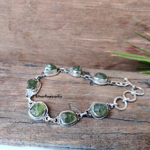 May include: A silver bracelet with a toggle clasp features seven green gemstone beads. The beads are irregularly shaped and have a rough, natural texture. The bracelet is on a wooden surface.