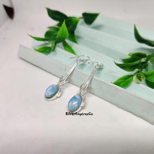 Könnte beinhalten: Ein Paar silberne Ohrringe mit einem hellblauen, ovalen Stein. Die Ohrringe haben ein langes, schlankes Design mit einem geschlungenen Akzent in der Nähe des Steins. Die Ohrringe werden auf einer hellblauen Oberfläche mit grünen Blättern präsentiert.