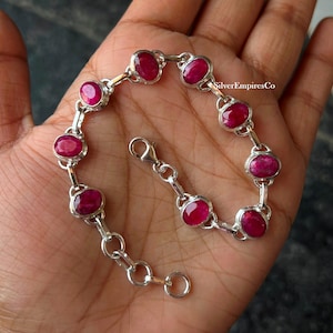 May include: A silver chain bracelet featuring eight oval-shaped, deep pinkish-red gemstones set in individual metal bezels. The bracelet includes a lobster claw clasp and a short extender chain with circular links. The text SilverEmpiresCo is visible.